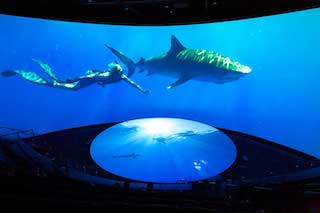 The new theatre has more than 300 seats and features an immersive 130-foot-wide by 32-foot-tall screen that curves in a 180-degree arc. A 30-foot-diameter disc tilts up from the floor to extend the projection surface